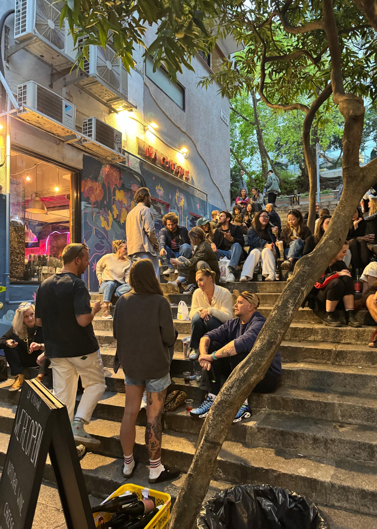 People sitting on outdoor steps in an urban setting with a store and trees in the background.
