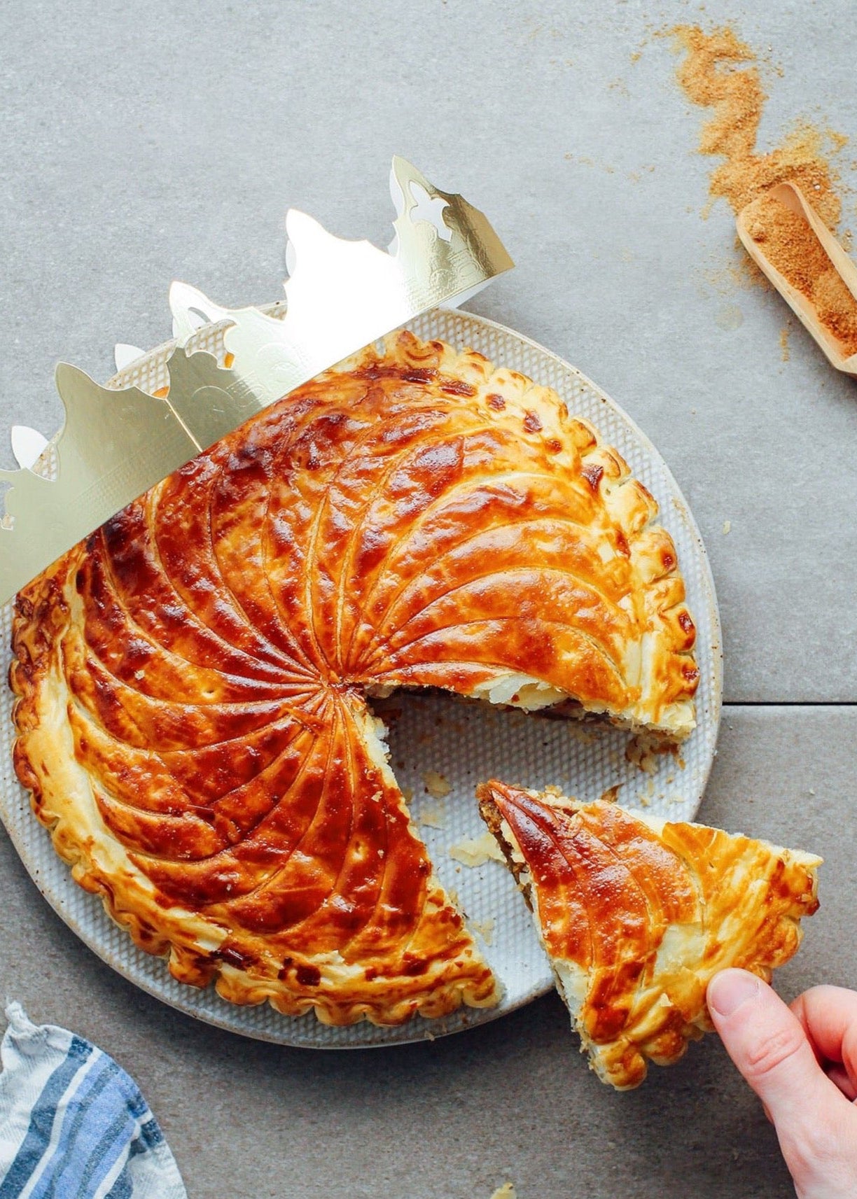 A traditional French Galette des Rois cake, with a creamy almond filling and flaky buttery layers, being sliced to reveal the filling. The cake is decorated with a golden, textured topping and a paper crown is visible on top.