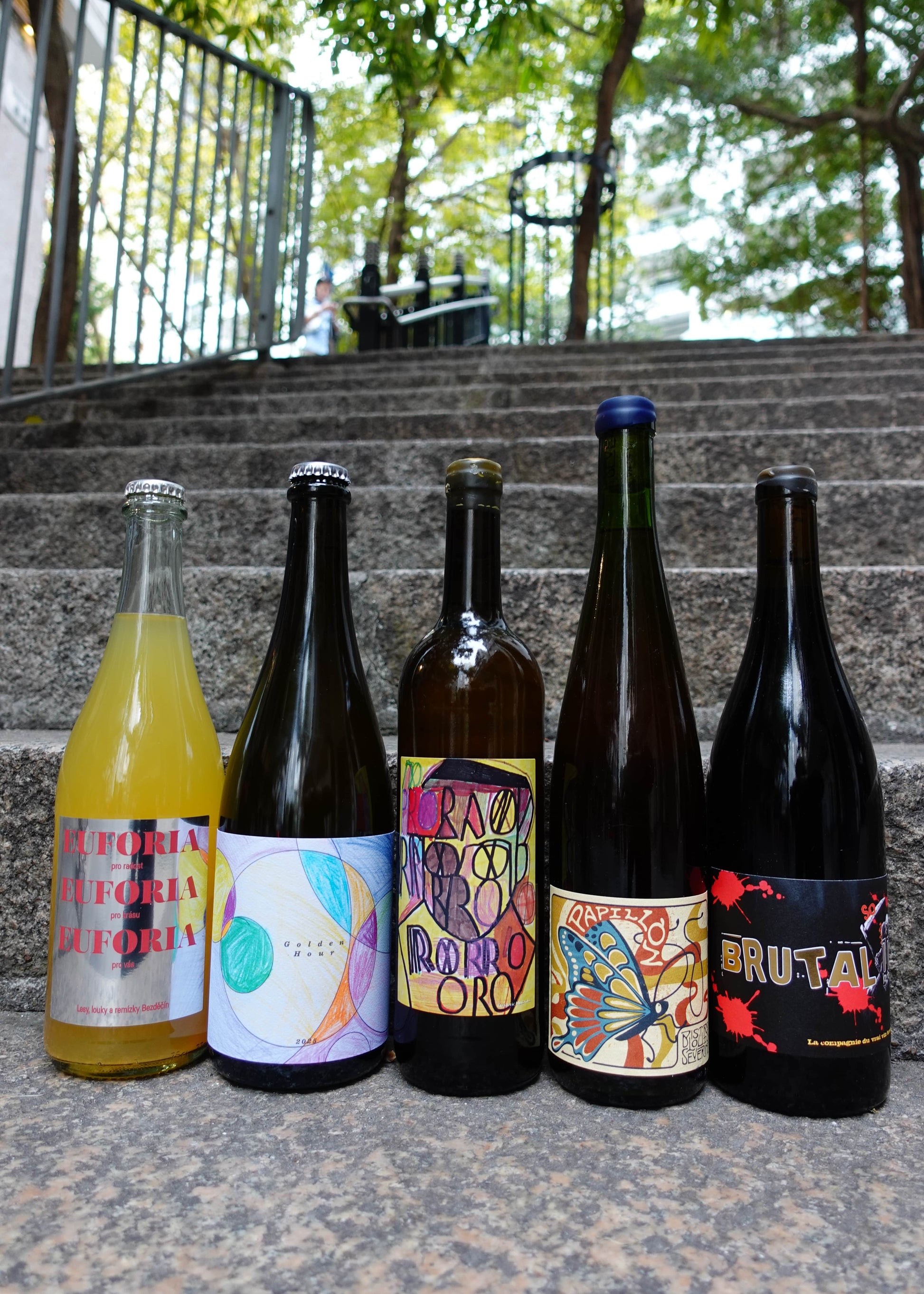 Five bottles of different types of drinks on a set of outdoor stairs with a blurred background.
